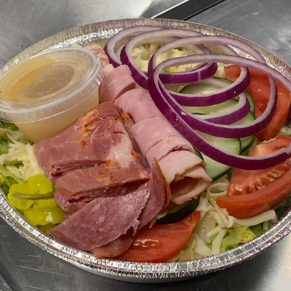 Antipasto salad with ham, salami, provolone, red onion, tomatoes, cucumbers, and dressing in a foil container.